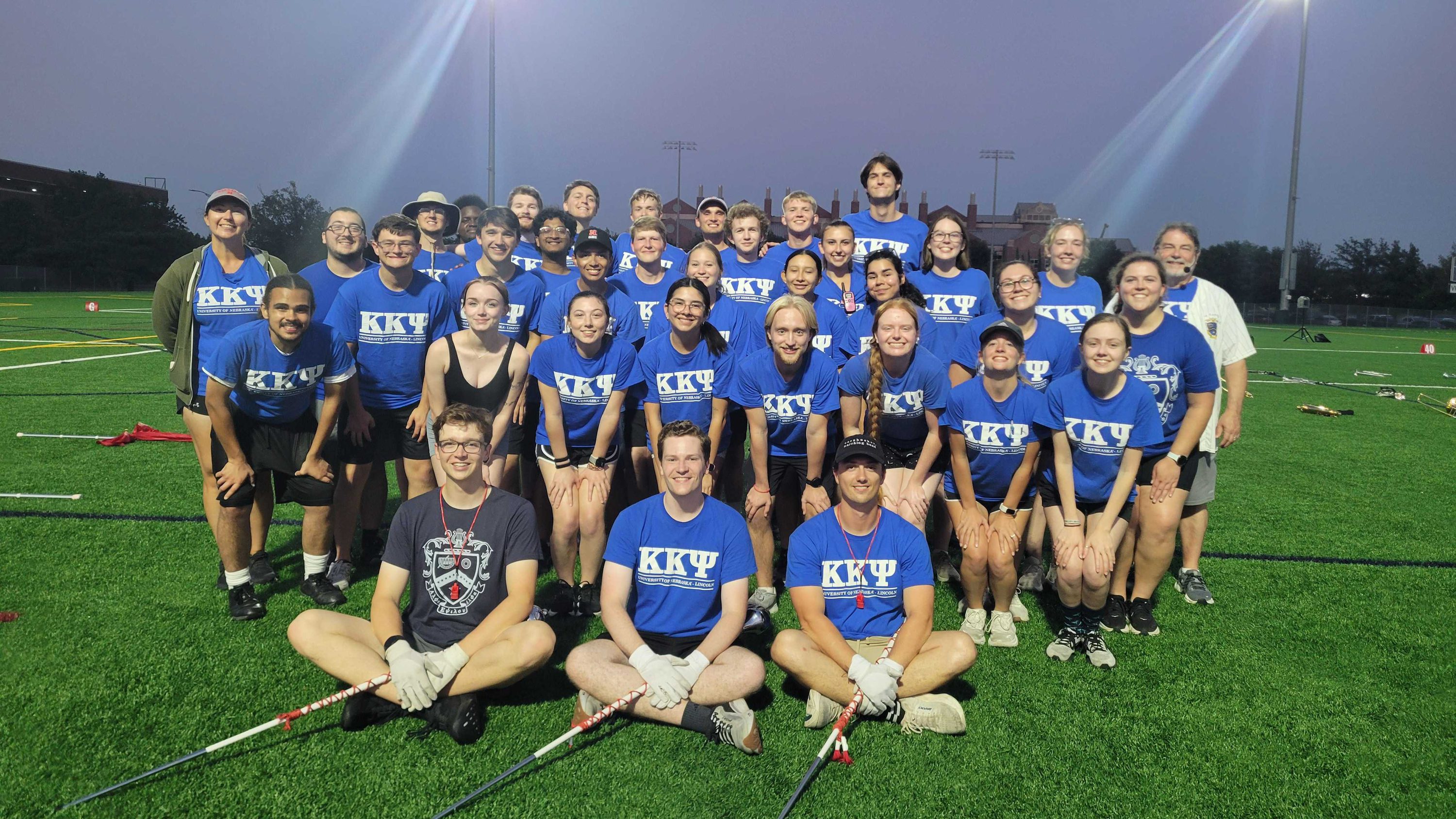 Group of people in blue KKY shirts posing outside on a lit sports field during dusk.